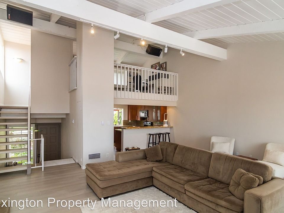 Living room with large brown sectional sofa, wooden floor, staircase, and kitchen in background