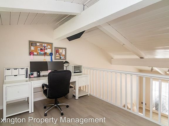 Home office with white desk, dual monitors, black chair, bulletin board, sloped ceilings, and wood floor