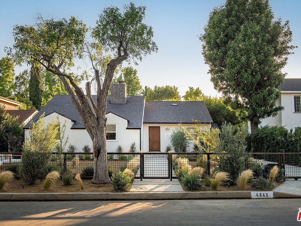 California real estate home with black roof, landscaped yard, trees, and front pathway