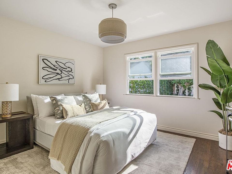 Bedroom with large bed, decorative pillows, nightstand with lamp, windows, plant, and abstract artwork