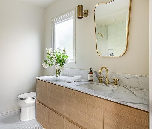 Bathroom with wooden vanity, marble countertop, round mirror, gold faucet, white toilet, and flowers