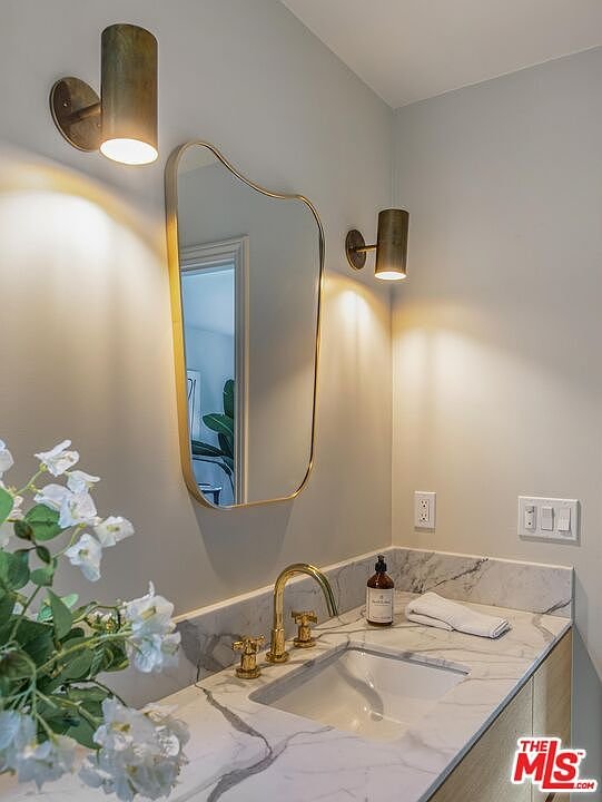 Bathroom with marble countertop, sink, large rounded mirror, wall lights, and decorative flowers
