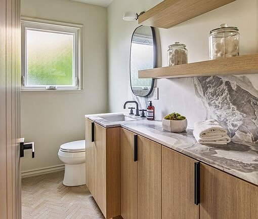 Bathroom with white toilet, wooden cabinets, marble countertop, oval mirror, and shelves with decor