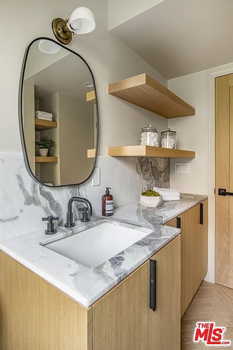 Bathroom with round mirror, sink on marble countertop, light wood cabinets, and shelves with decor