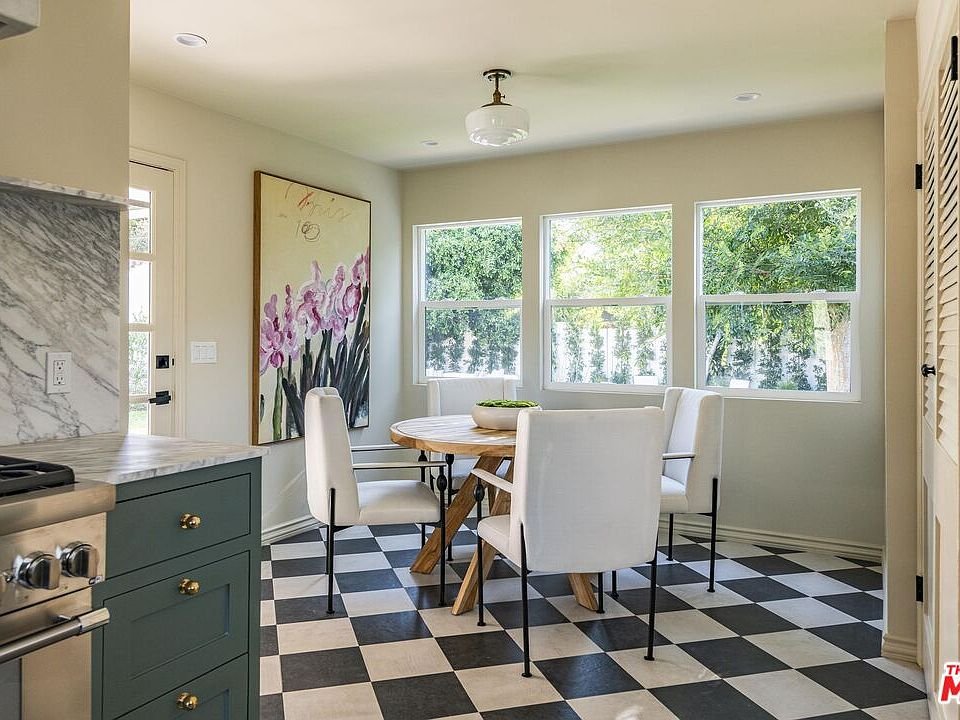 Kitchen dining area with round table, white chairs, large windows, pendant light, black and white tile floor, and floral artwork