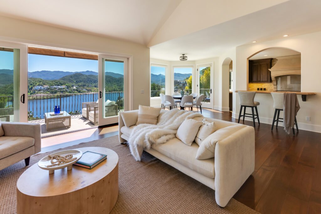 Living room with sofa, round wooden coffee table, large windows, mountain view, and kitchen bar.