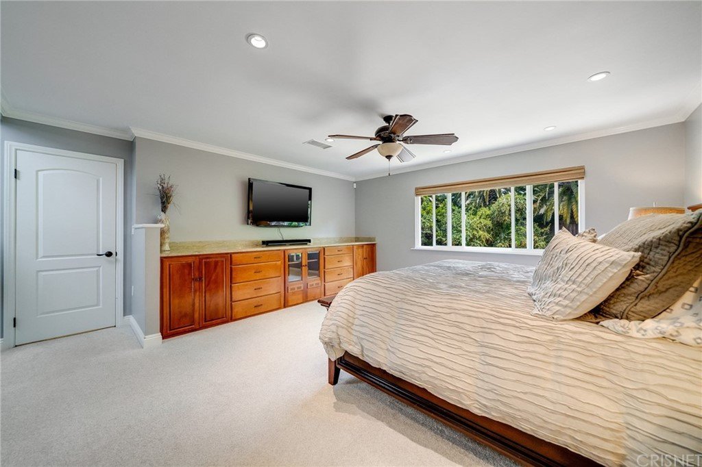 Bedroom with wood bed, light bedding, dresser, wall TV, window, and carpet floor.