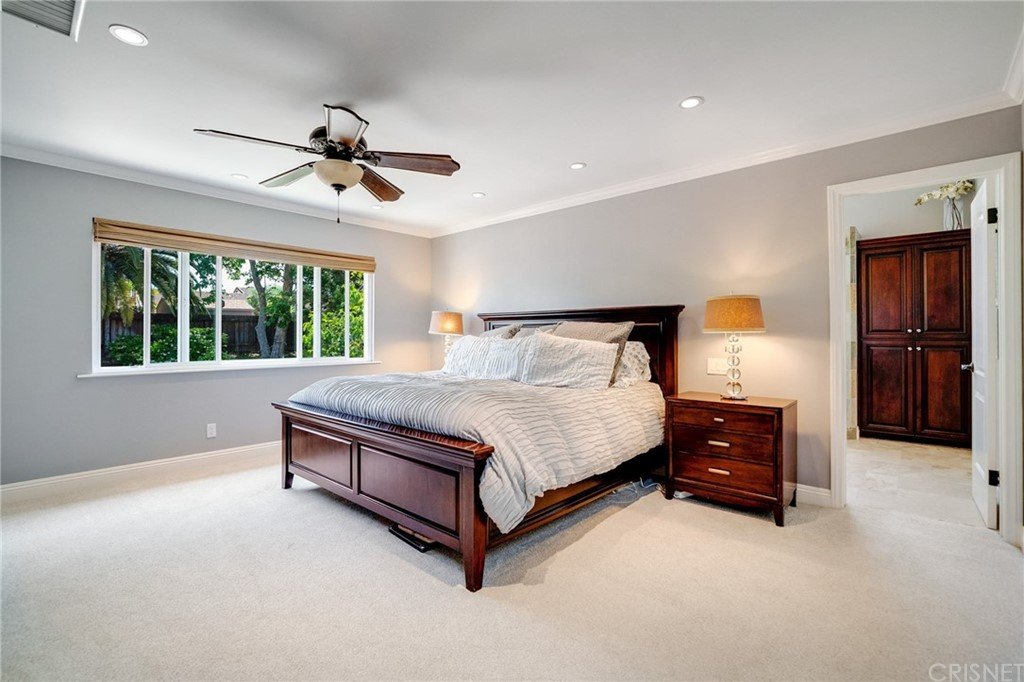 Bedroom with bed, white linens, wood nightstands, ceiling fan, windows, and green view.