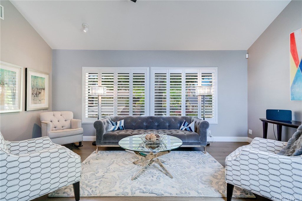 Living room with gray sofa, two chairs, glass coffee table, rug, window shutters, and pictures on the wall.