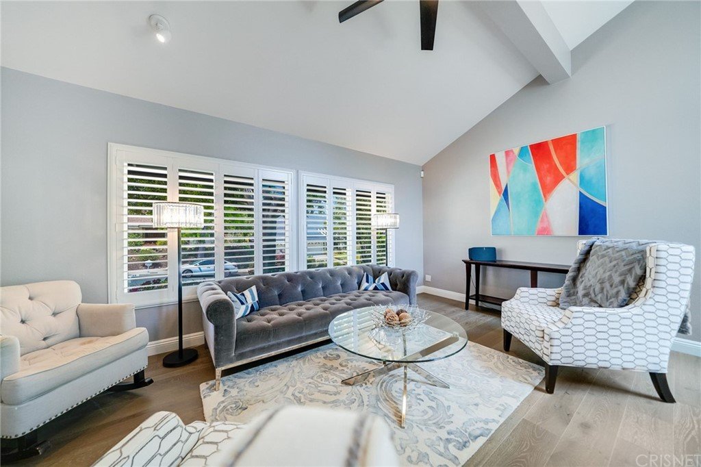 Living room with gray sofa, white chairs, glass coffee table, rug, large windows, and wall art.
