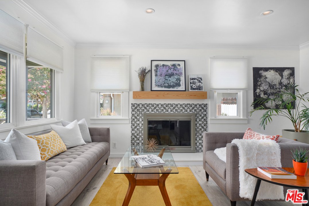 Living room with two gray sofas, orange rug, glass coffee table, fireplace, large windows, and wall art.