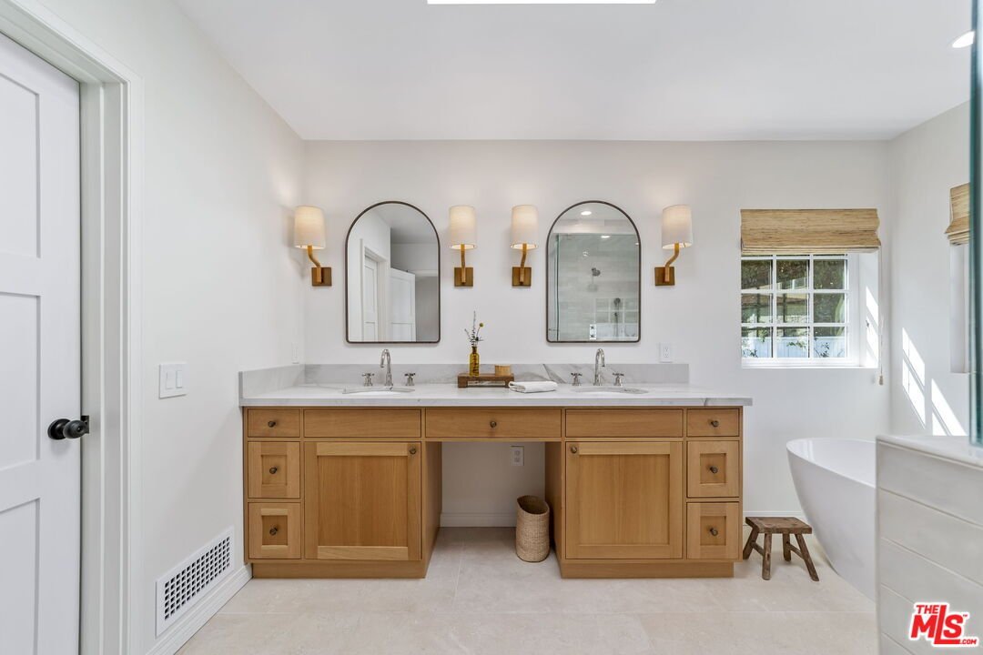 Modern bathroom with double sinks, mirrors, wood cabinets, bath tub, and window.