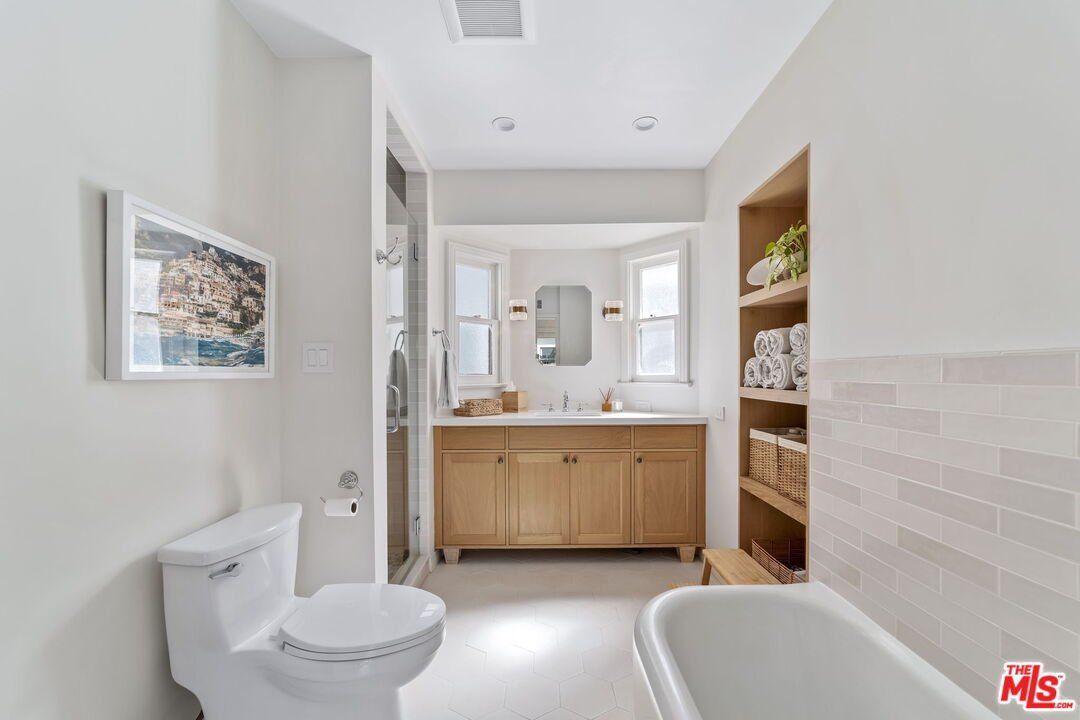 Modern bathroom with white bath tub, beige vanity, mirror, shelves, towels, and window.