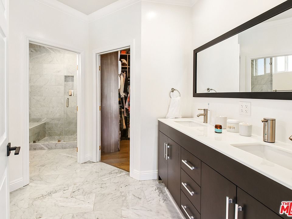 Bathroom with double sink vanity, mirror, glass shower, marble floor, and door to closet.