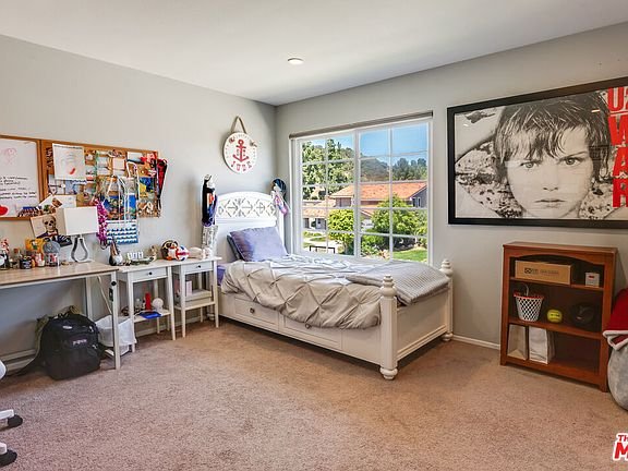 Bedroom with bed, desk with supplies, bulletin board, bookshelf, wall art, big windows, and carpet floor.