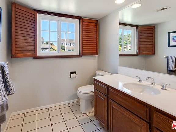 Bathroom with double sink vanity, wood cabinets, white toilet, window with shutters, and tile floor.
