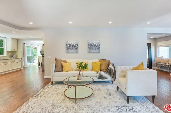 Living room with white sofa, pillows, glass coffee table, vase with flowers, wood floor, and wall art.
