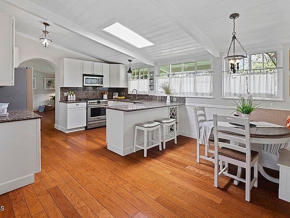 Kitchen with white cabinets, granite counter, steel appliances, round table with chairs, wood floor, and big windows.