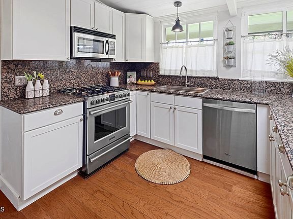 Kitchen with white cabinets, steel oven and microwave, granite counter, sink under window, rug, and wood floor.