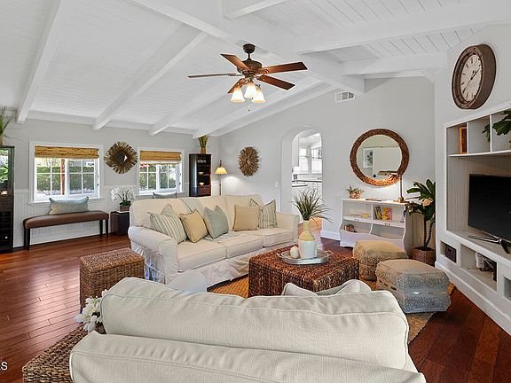 Living room with white sectional sofa, pillows, wood coffee table, baskets, plants, wall TV, shelves, big windows, and ceiling fan.