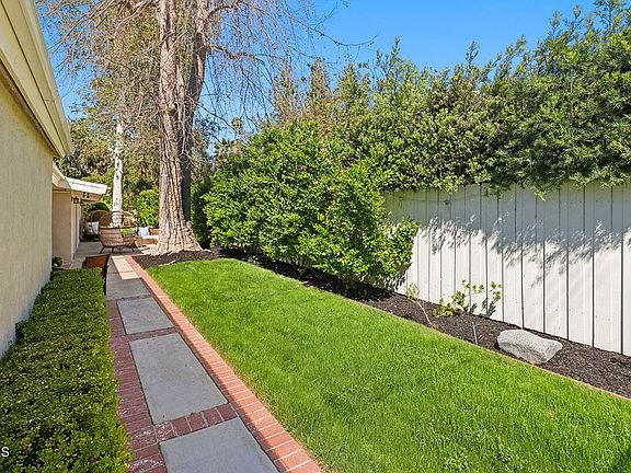 Garden path with grass and green plants beside a house with white fence California real estate.