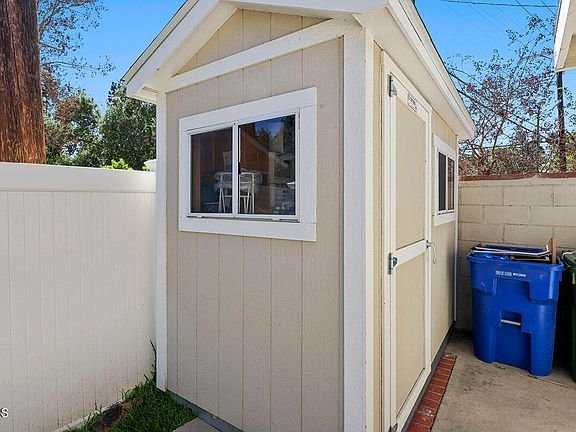 Beige storage shed with white trim, windows, white fence, and blue trash bin nearby.