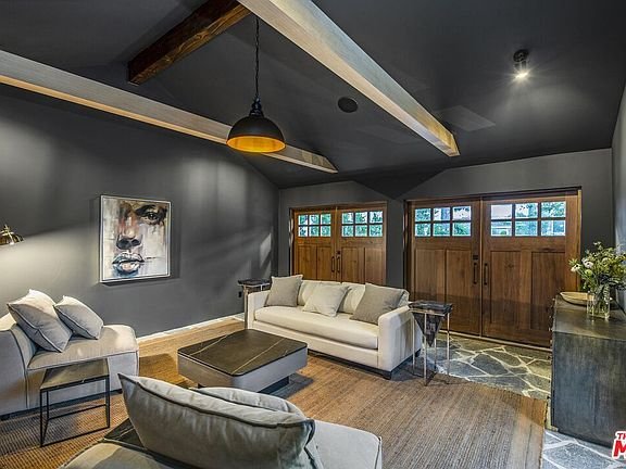 Living room with dark wall, wooden beams, large face artwork, light sofas, and double wooden doors